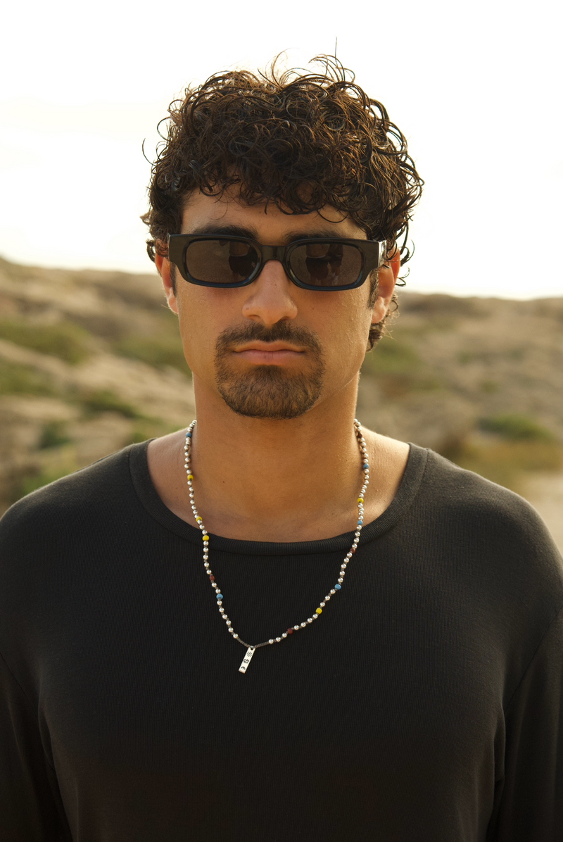 Man wearing sunglasses and a black shirt with a necklace, standing against a natural landscape.